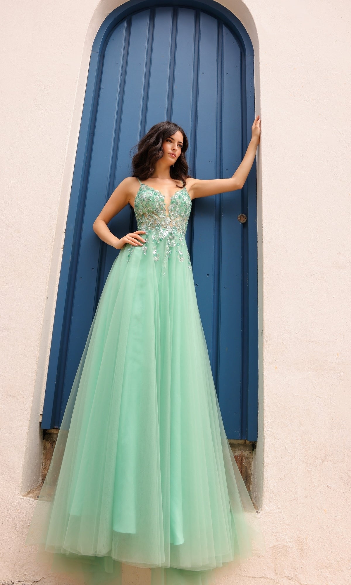 A woman in the Deep V-Back Long Tulle A-Line Prom Dress Q1391, featuring embroidered flowers, stands before a tall arched blue door set in a pale wall.